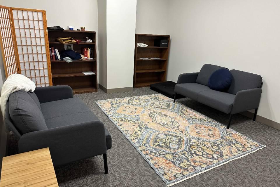 A room with sofas along each wall and a rug in the center. There are book shelves in the background with various items visible.