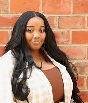 Aubra Ladd, headshot, stands in front of a brick wall and smiles. She is wearing a tan and white plaid shirt over a brown top.