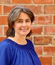 Leah Sweetman, headshot, wears a blue blouse and stands in front of a red brick wall, smiling.